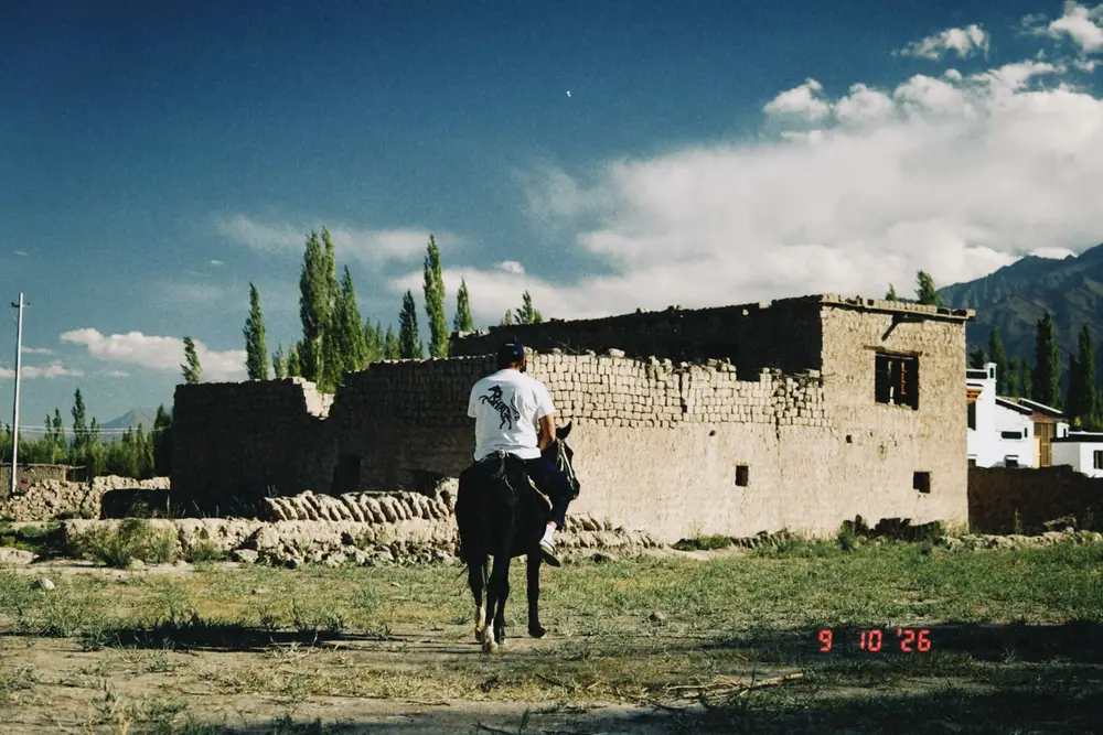 A rider in a white BHAG horse-print tee riding past a mud-brick house in Ladakh